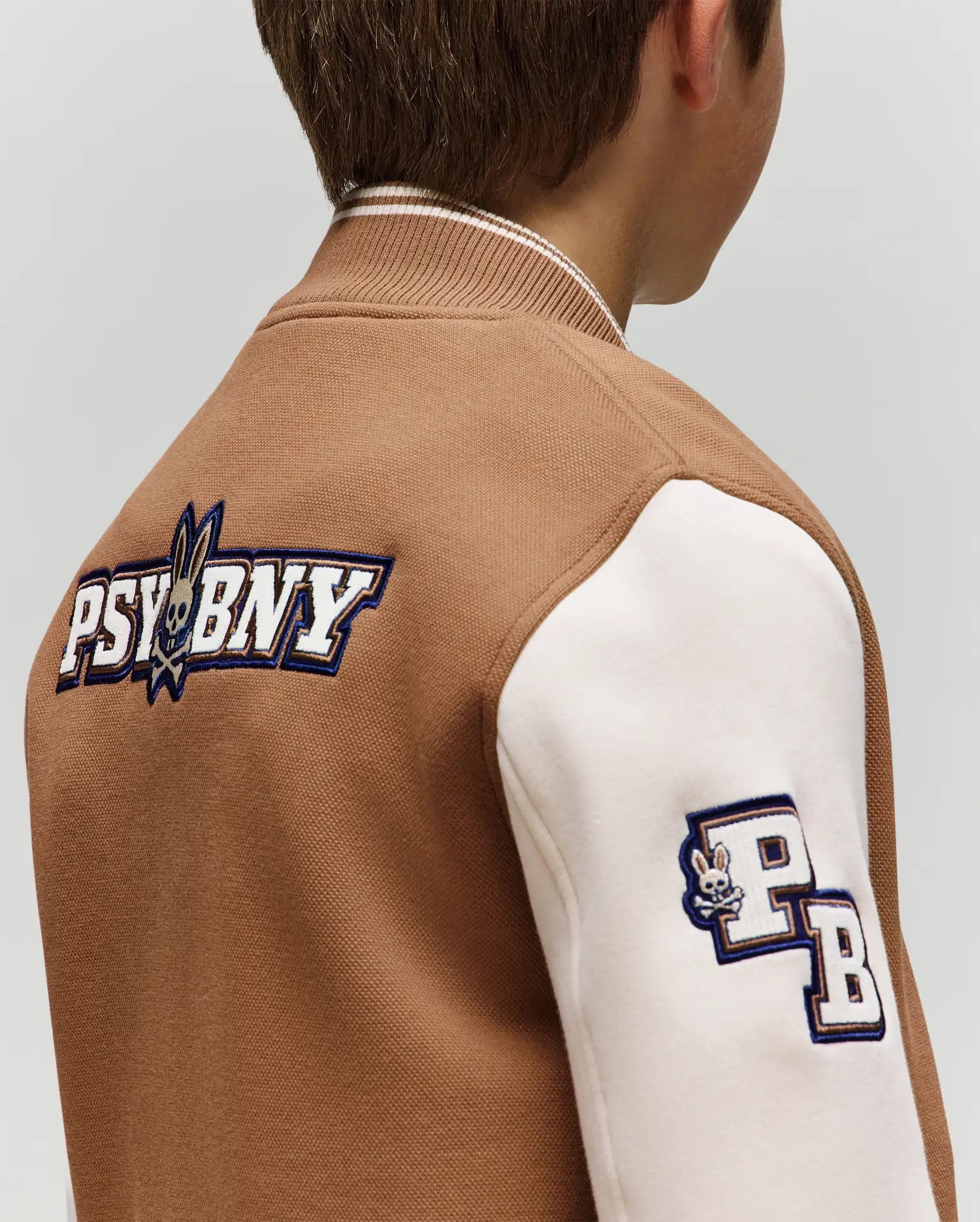 A child wears the Psycho Bunny KIDS WADE KNIT BOMBER (KO1100001), a brown and white varsity jacket with “PSYBNY” and a bunny logo on the back, plus a “PB” patch with a small bunny head on the sleeve. Seen from behind.