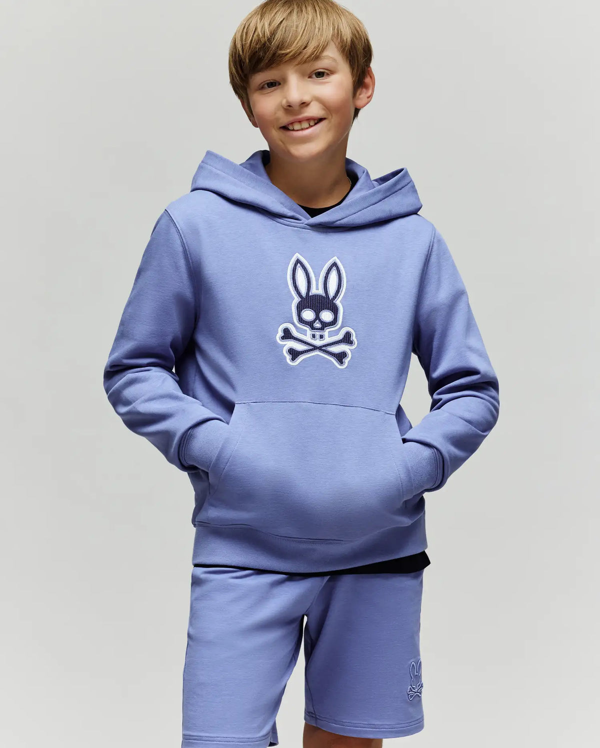 A smiling boy with light brown hair wears the Psycho Bunny KIDS RYDER BUNNY HOODIE (KT0500018), featuring an embroidered Bunny patch, standing hands-in-pockets against a plain background—the perfect loungewear look.