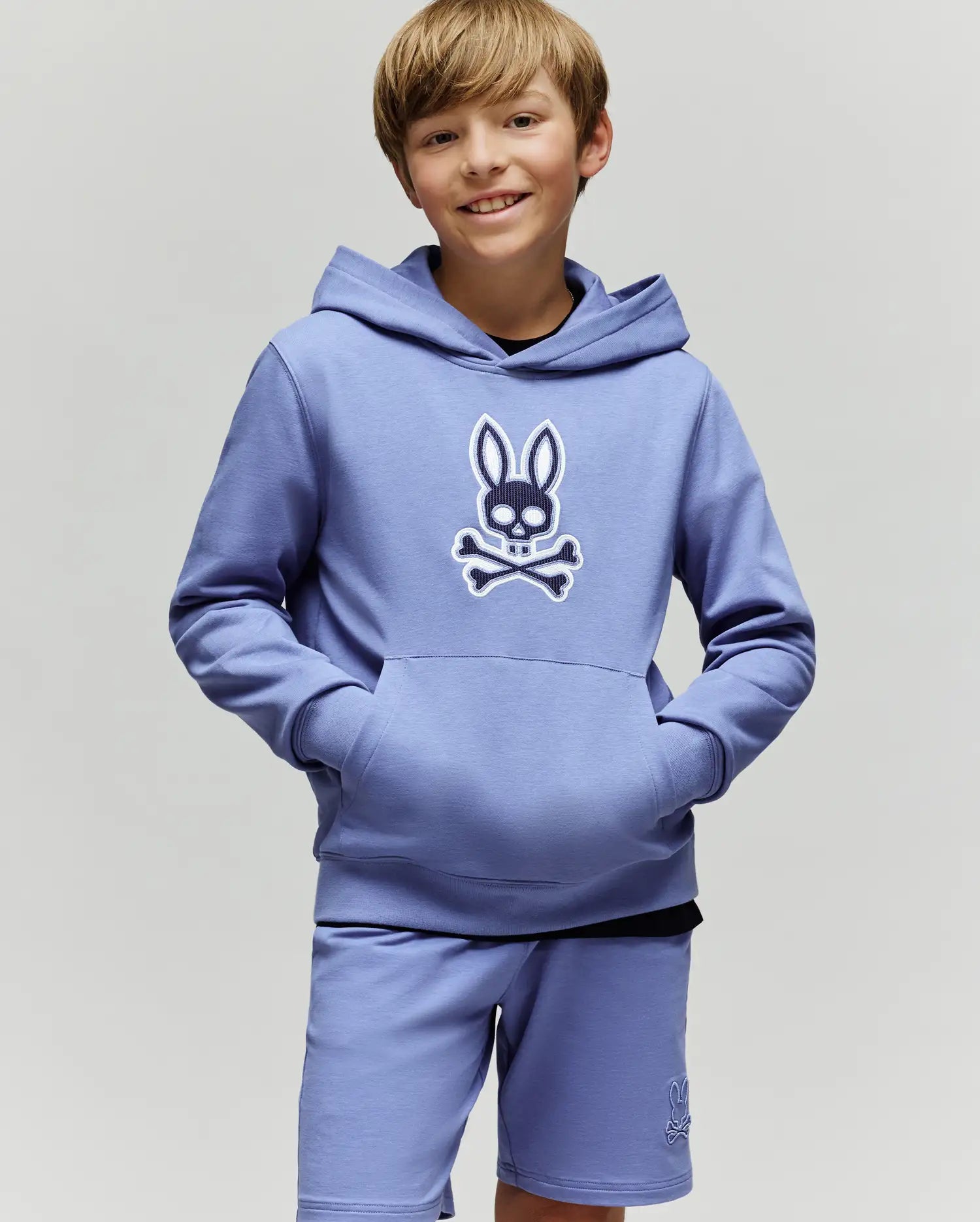 A smiling boy with light brown hair wears the Psycho Bunny KIDS RYDER BUNNY HOODIE (KT0500018), featuring an embroidered Bunny patch, standing hands-in-pockets against a plain background—the perfect loungewear look.