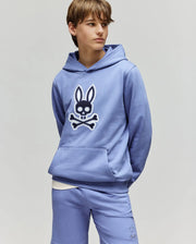 A teen boy stands with hands in pockets, wearing the Psycho Bunny KIDS RYDER BUNNY HOODIE (KT0500018) in light blue with an embroidered Bunny patch, matching shorts, and a white shirt underneath against a plain background.