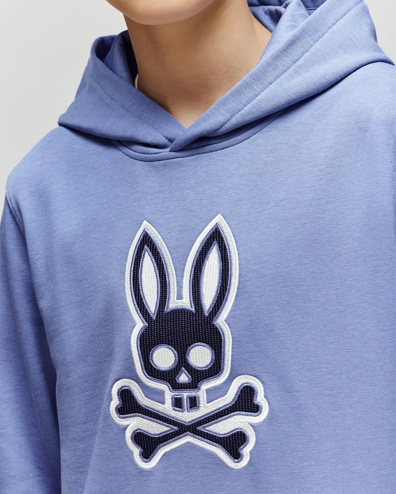A person wears the Psycho Bunny KIDS RYDER BUNNY HOODIE (KT0500018) in light blue, featuring a large embroidered Bunny patch. The cropped image shows only the upper torso and part of the face, ideal for loungewear inspiration.