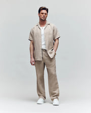 A man wears Psycho Bunny’s MENS FARRIS WIDE LEG LINEN PANT (MB0600007) in light brown with a matching short-sleeve shirt over a white tee, paired with white sneakers and sunglasses, standing against a light background with one hand in his pocket.