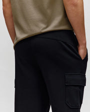 A person in Psycho Bunny’s MENS NASH CARGO SWEATPANT (MB0800010)—black cargo sweatpants with side pockets—stands with one hand in their pocket, waist-level from the back, blending comfort and practical style.