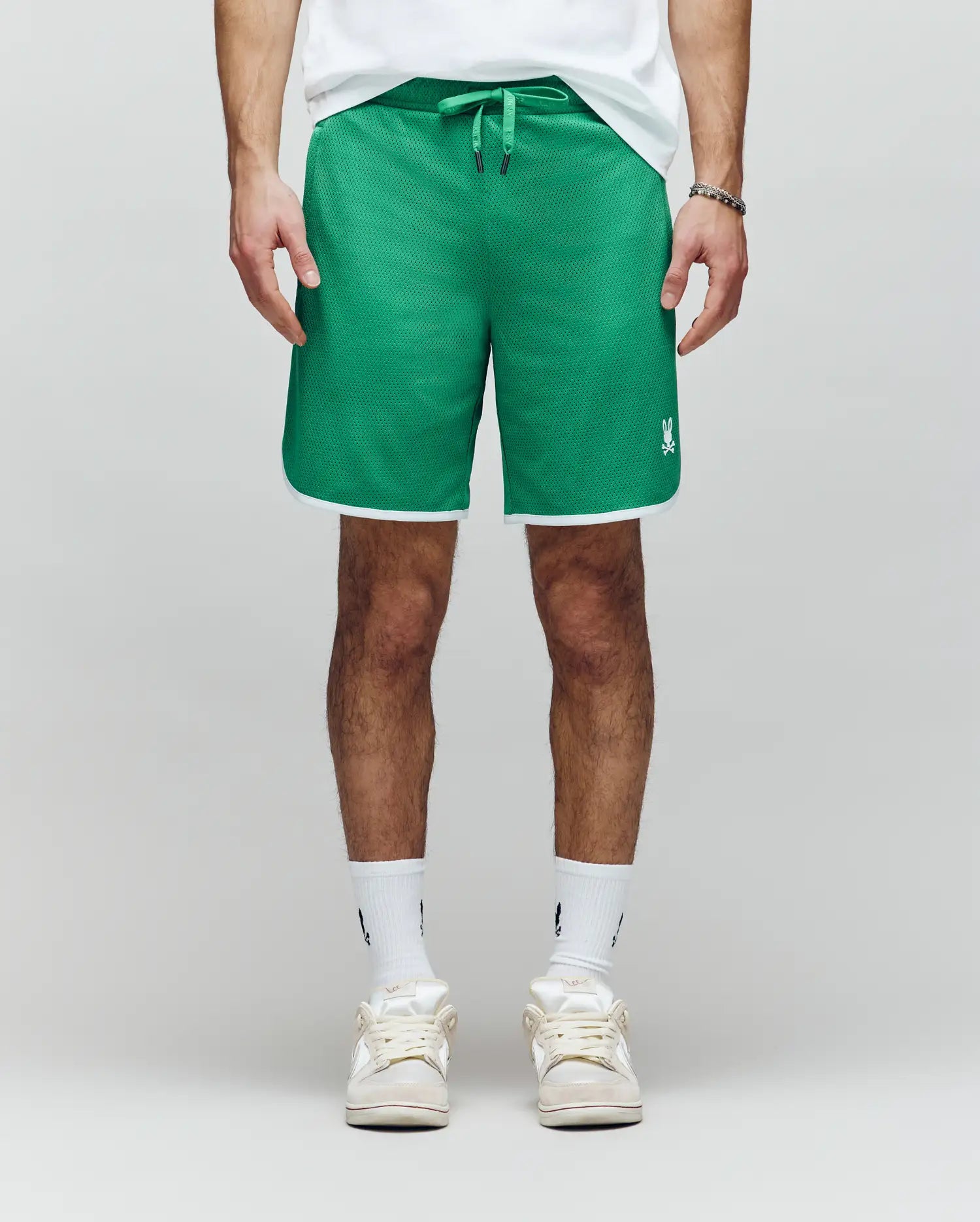 A person wearing Psycho Bunny MENS NICO MESH SHORT - MB0900004 in green with white trim, a white T-shirt, white crew socks, and white sneakers stands against a light gray background.