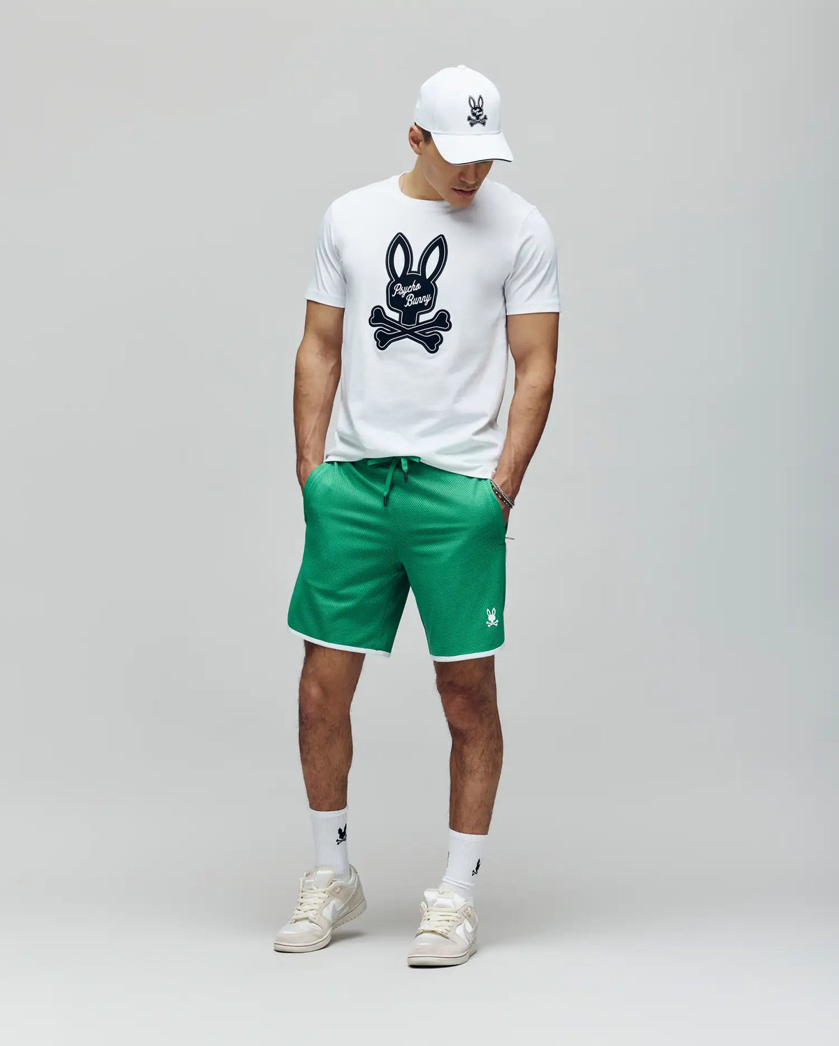 A man stands against a plain background wearing Psycho Bunny's MENS NICO MESH SHORT - MB0900004, a white cap, a white graphic T-shirt with the bunny logo, and breathable mesh sneakers, hands in pockets for effortless athletic comfort.