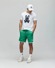A man stands against a plain background wearing Psycho Bunny's MENS NICO MESH SHORT - MB0900004, a white cap, a white graphic T-shirt with the bunny logo, and breathable mesh sneakers, hands in pockets for effortless athletic comfort.