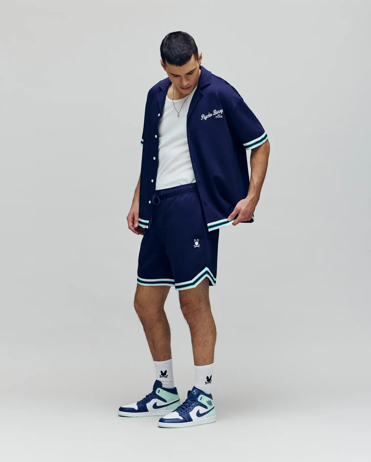 A man stands looking down, wearing a navy short-sleeve button-up shirt, the Psycho Bunny MENS BASIL TAPED SHORT - MB0900005 with an elastic waistband and light blue trim, a white tank top, white socks, and blue and white sneakers against a plain background.