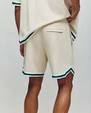 A person stands with their back to the camera wearing Psycho Bunny's MENS BASIL TAPED SHORT - MB0900005 in premium cotton-modal, featuring green and blue striped trim and an embroidered bunny, against a plain light gray background.