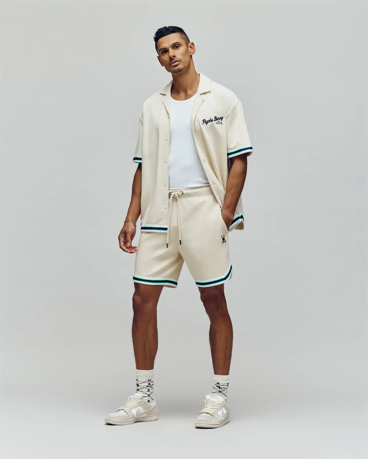 A man stands against a plain background wearing Psycho Bunny MENS BASIL TAPED SHORT - MB0900005 with green and black trim, a cream cotton-modal short-sleeve shirt, a white tank top, patterned socks, and white high-top sneakers.