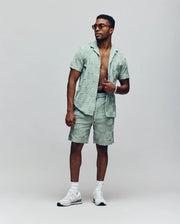 A man stands confidently against a plain background in Psycho Bunny's Kelvin Towel Terry Sweatshort (MB0900006) paired with a light green jacquard terry cloth short-sleeve shirt, open to reveal his chest, white sneakers, and sunglasses.