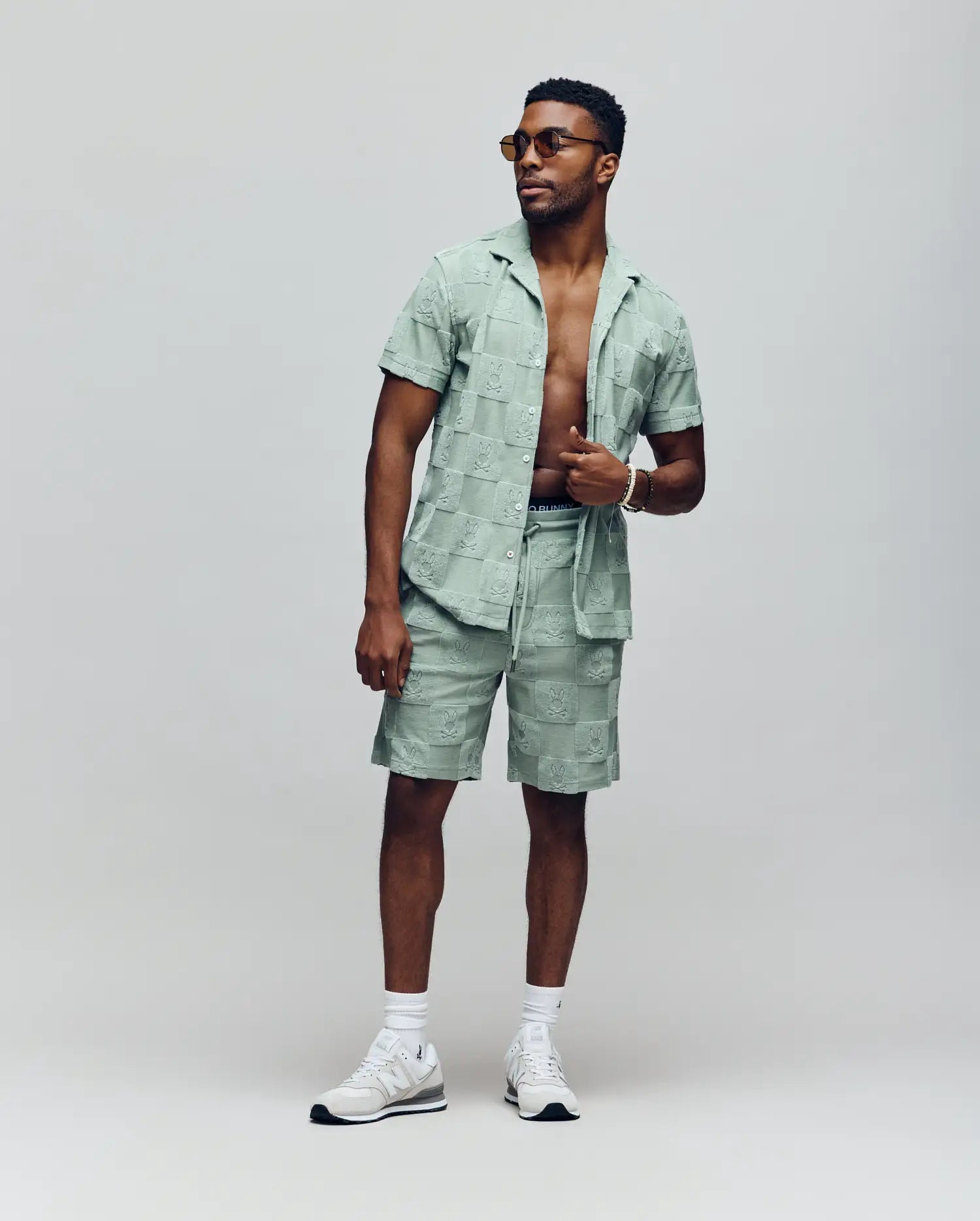 A man stands confidently against a plain background in Psycho Bunny's Kelvin Towel Terry Sweatshort (MB0900006) paired with a light green jacquard terry cloth short-sleeve shirt, open to reveal his chest, white sneakers, and sunglasses.