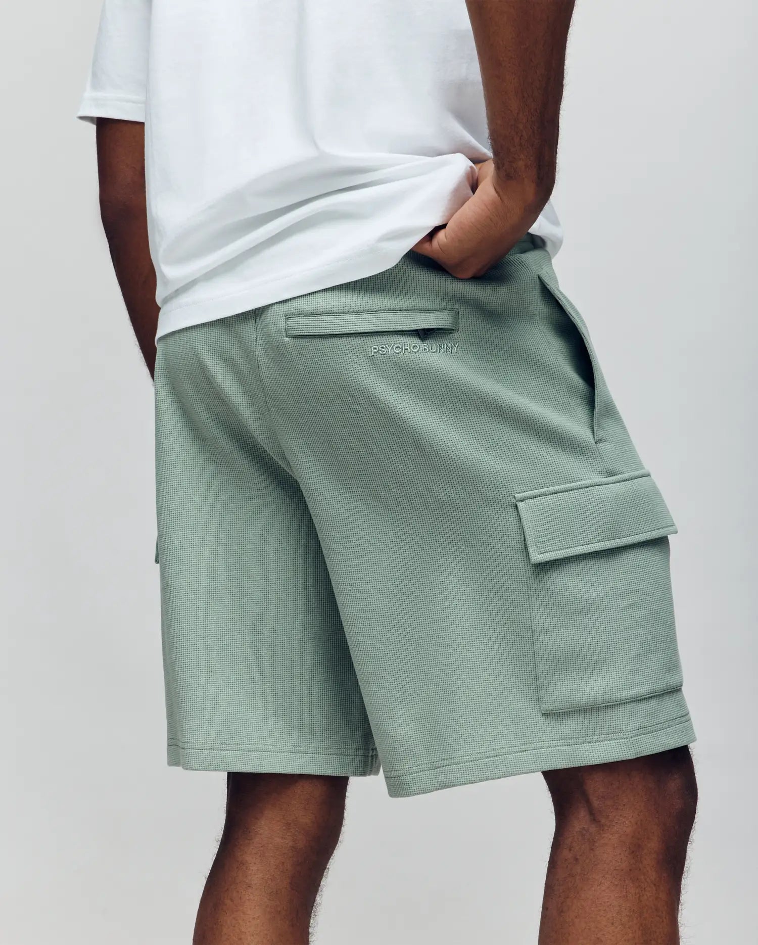 A person is seen from the back wearing a white waffle knit tee and Psycho Bunny MENS RON WAFFLE CARGO SHORT - MB0900012 in light green, featuring side and back pockets, embroidered logos, relaxed fit, and one hand in a pocket.