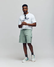 A man stands against a plain background wearing a white graphic tee, gray socks, green and white sneakers, and Psycho Bunny’s MENS RON WAFFLE CARGO SHORT (MB0900012) in light green with embroidered logos. He holds sunglasses confidently.