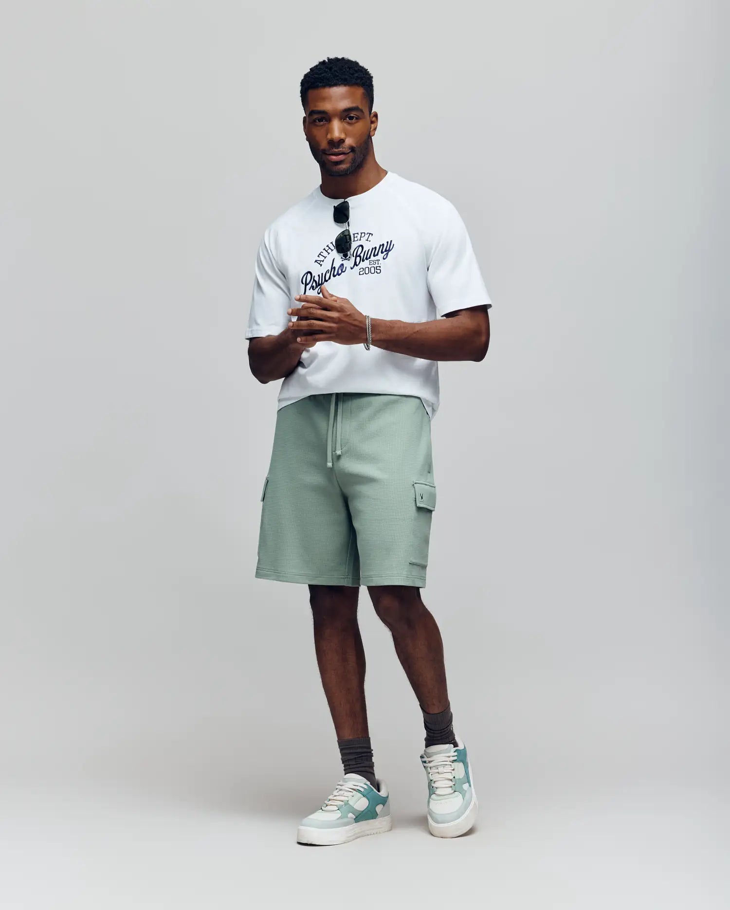 A man stands against a plain background wearing a white graphic tee, gray socks, green and white sneakers, and Psycho Bunny’s MENS RON WAFFLE CARGO SHORT (MB0900012) in light green with embroidered logos. He holds sunglasses confidently.