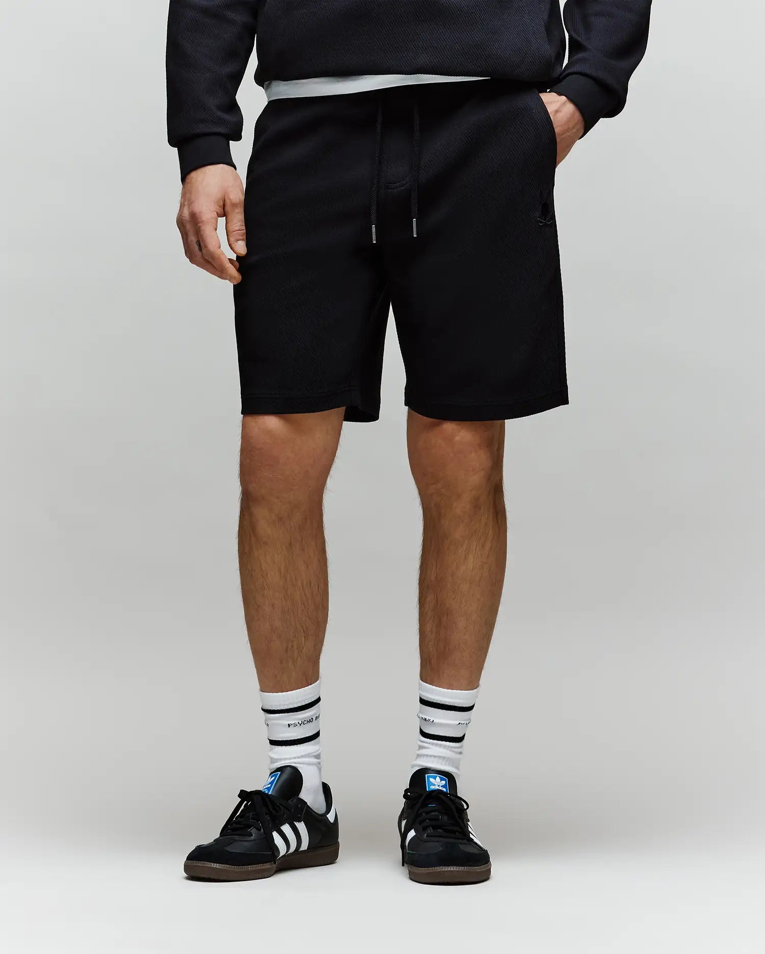 A person in a black sweatshirt, Psycho Bunny MENS KAI SWEATSHORT - MB0900015 featuring an embroidered Bunny logo, white socks with black stripes, and black sneakers with white stripes stands against a plain light background.