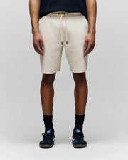 A person wearing Psycho Bunny MENS KAI SWEATSHORT - MB0900015 in beige with an embroidered Bunny logo, a black T-shirt, black crew socks, and black sneakers stands against a plain light gray background.
