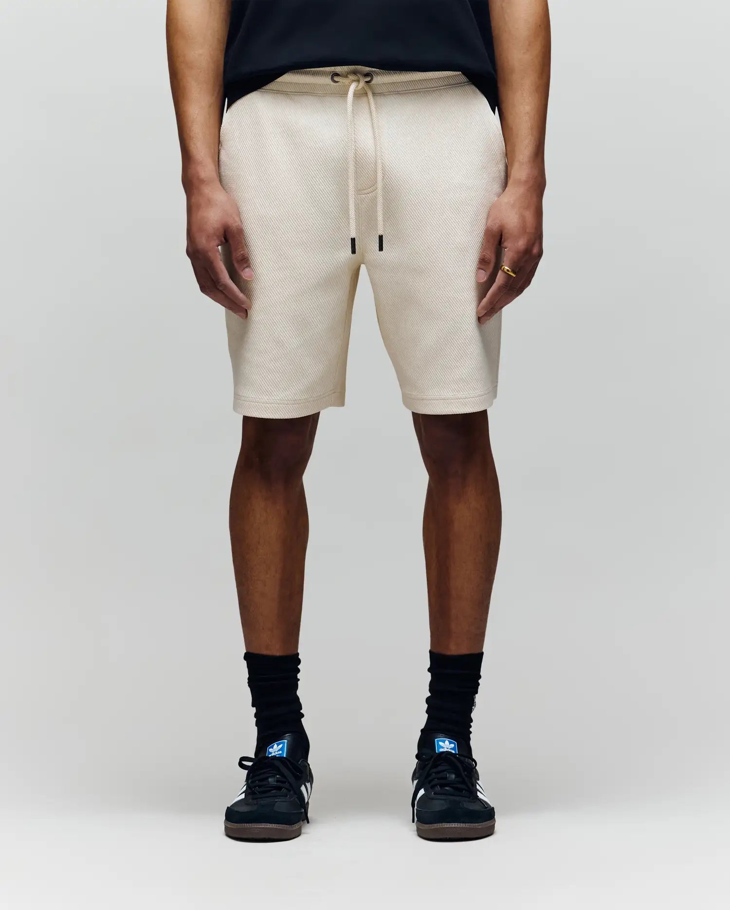 A person wearing Psycho Bunny MENS KAI SWEATSHORT - MB0900015 in beige with an embroidered Bunny logo, a black T-shirt, black crew socks, and black sneakers stands against a plain light gray background.