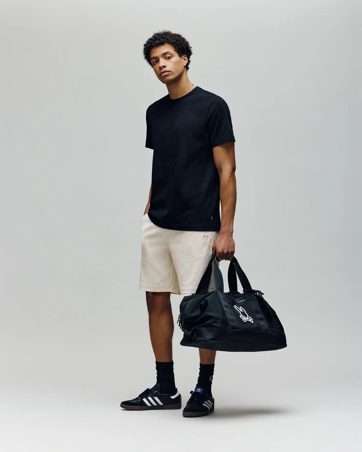 A young man stands against a plain background in a black T-shirt, Psycho Bunny MENS KAI SWEATSHORT (MB0900015) with embroidered Bunny logo, black socks, black Adidas sneakers, and holds a large black duffel bag, looking relaxed.