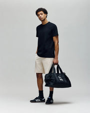A young man stands against a plain background in a black T-shirt, Psycho Bunny MENS KAI SWEATSHORT (MB0900015) with embroidered Bunny logo, black socks, black Adidas sneakers, and holds a large black duffel bag, looking relaxed.