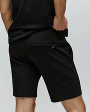 A person stands with one hand in their pocket, wearing Psycho Bunny’s MENS LAZSLO SWEATSHORT - MB0900016 in black, featuring a stretch modal-cotton blend and zippered back pocket, ideal for casual looks against a light background.