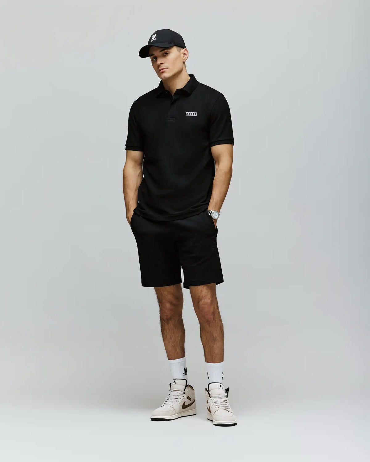 A man in a black polo, Psycho Bunny MENS LAZSLO SWEATSHORT - MB0900016, white socks and sneakers, and a black cap stands hands-in-pockets against a light gray background for a perfected casual look.