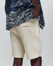 A person wears Psycho Bunny MENS LAZSLO SWEATSHORT - MB0900016 in a stretch modal-cotton blend with side and back pockets, paired with a navy cityscape-patterned shirt. The photo shows the look from waist to knees.