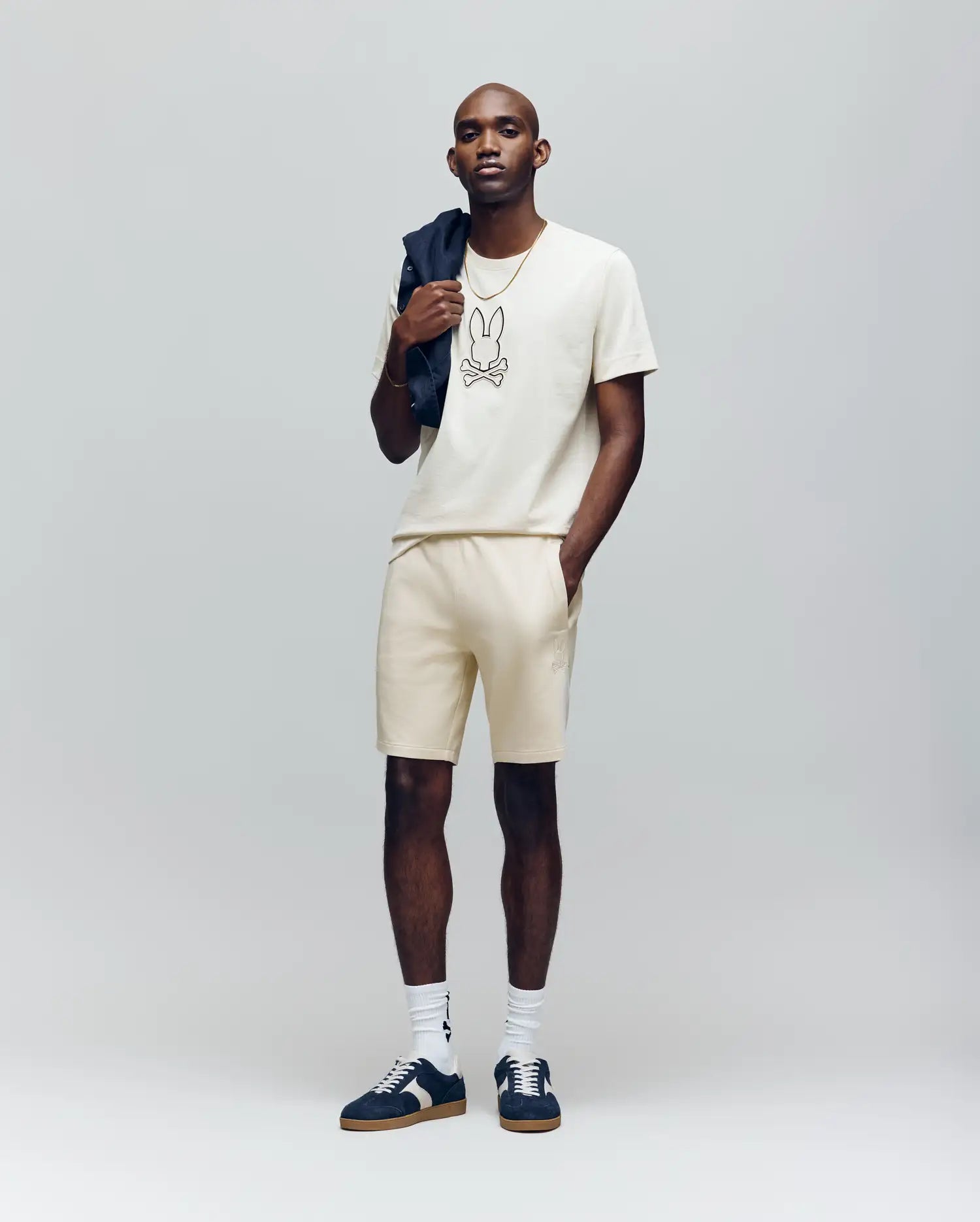 A man stands against a plain background wearing a cream stretch modal-cotton-blend T-shirt with an embroidered Bunny, Psycho Bunny MENS LAZSLO SWEATSHORT MB0900016, white socks, black sneakers, and holding a dark jacket over his shoulder.