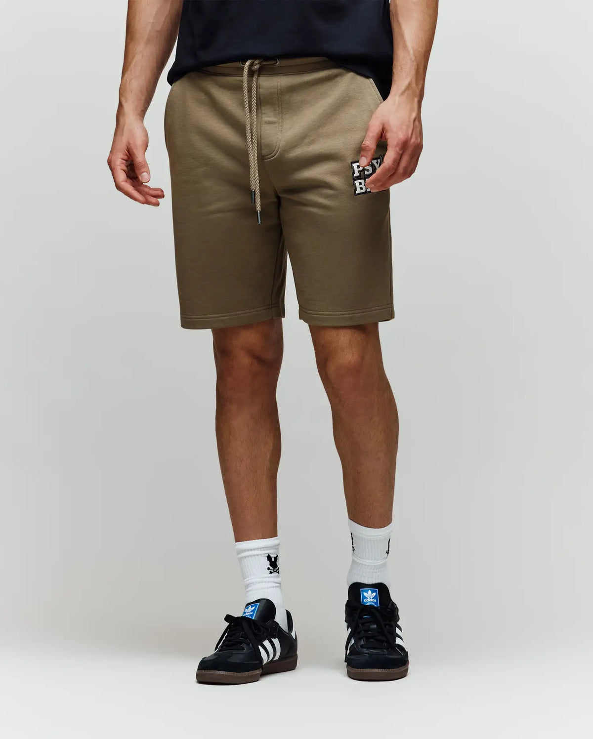A person wearing Psycho Bunny MENS WADE SWEATSHORT - MB0900028 in tan, featuring an embroidered “FEVR” wordmark, paired with white socks and black Adidas sneakers, stands against a plain light background.