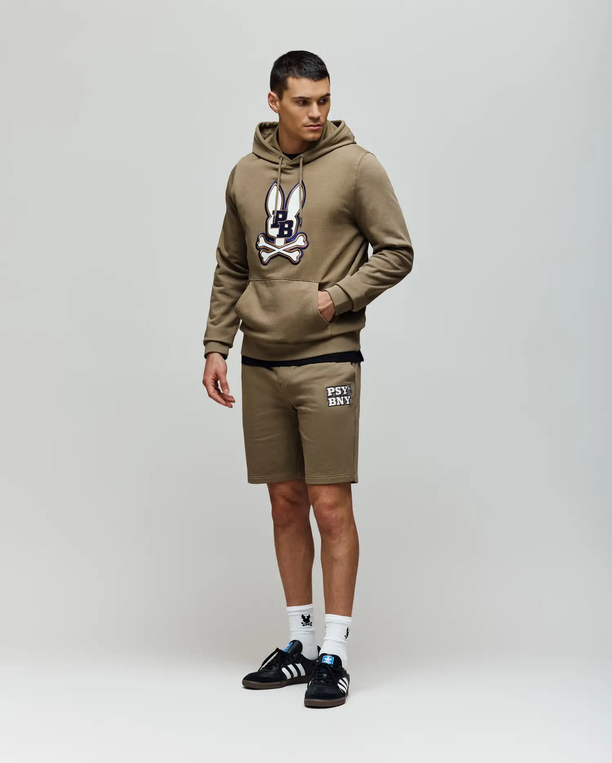 A man in Psycho Bunny MENS WADE SWEATSHORT - MB0900028 poses against a plain background, wearing an olive green hoodie, olive shorts with white text, white socks, and black Adidas sneakers for a classic athletic look.