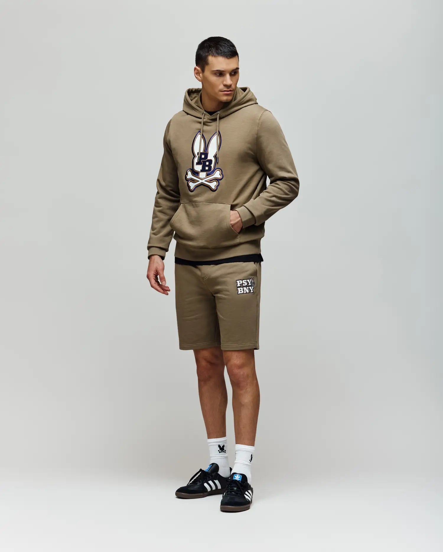 A man in Psycho Bunny MENS WADE SWEATSHORT - MB0900028 poses against a plain background, wearing an olive green hoodie, olive shorts with white text, white socks, and black Adidas sneakers for a classic athletic look.