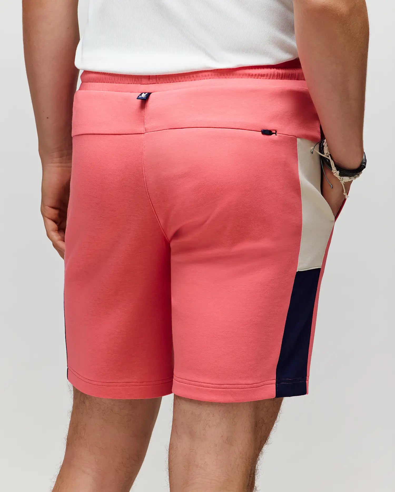 A person wears the Psycho Bunny MENS DAWSON TRACK SHORT - MB0900032 in pink with navy and beige side panels, styled with a white shirt and wristwatch for a relaxed street style vibe, photographed from behind.
