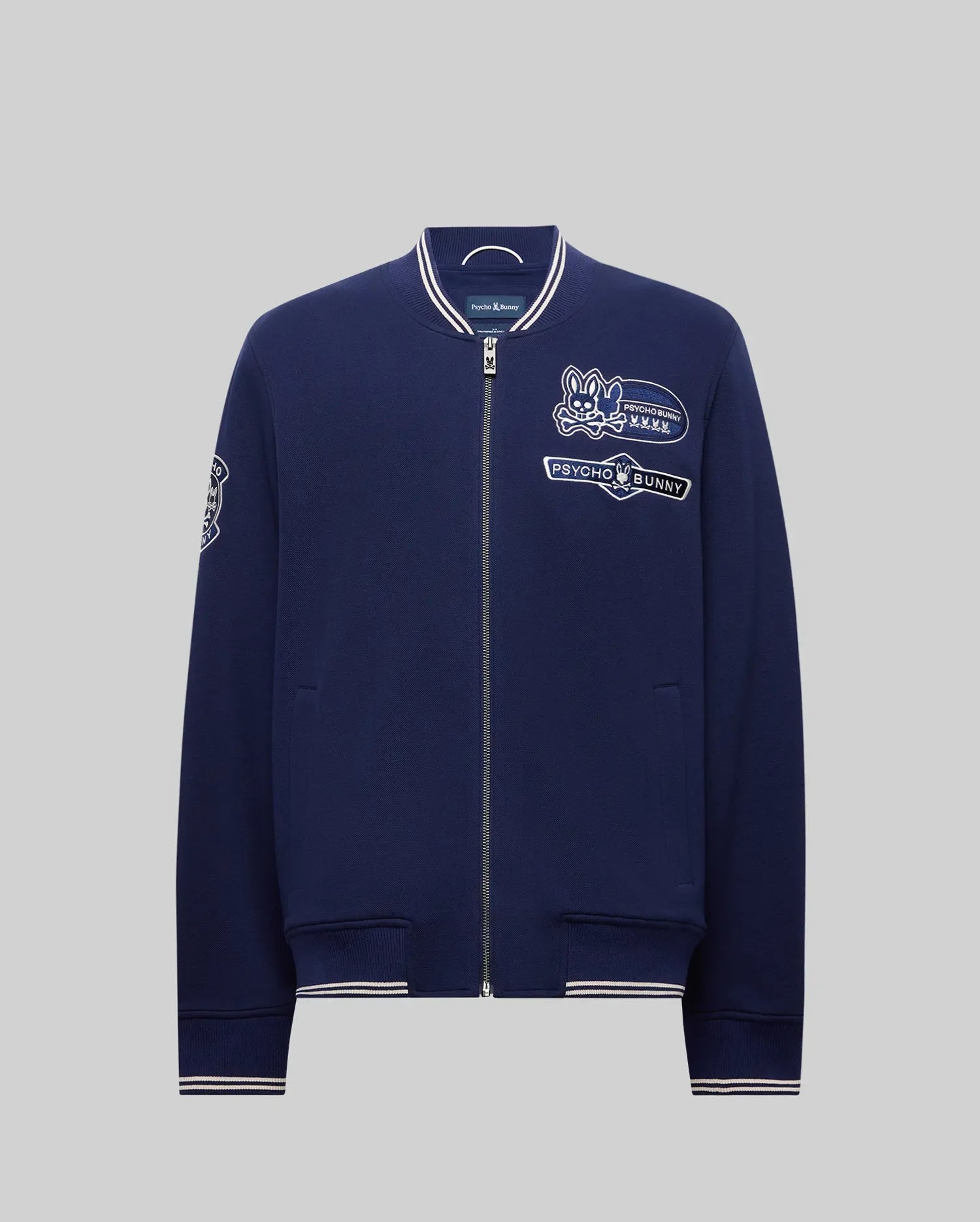 The Psycho Bunny MENS DESMOND PIQUE KNIT BOMBER - MO1100018 is a navy bomber with white trim, varsity-style details, and embroidered patches including a bunny logo, "Psycho Bunny" text on the chest, and a sleeve patch.
