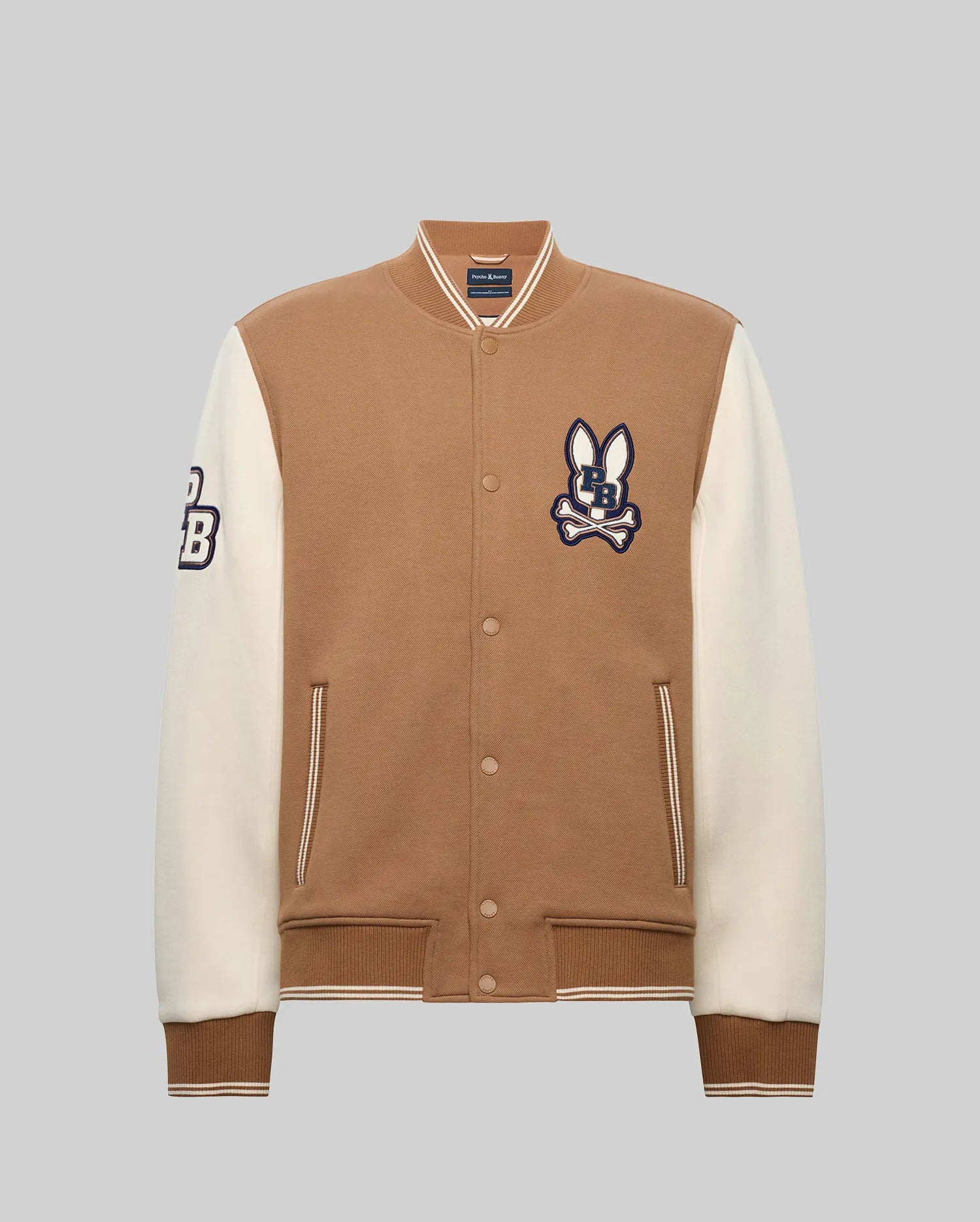 The Psycho Bunny MEN'S WADE KNIT BOMBER (MO1100020) features a tan body, white sleeves, an embroidered bunny "CB" logo on the chest, a "CB" patch on the sleeve, front pockets, and ribbed cuffs.