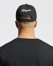 A person is shown from behind wearing a Psycho Bunny MENS LOS ANGELES BASEBALL CAP (MO1500013) with "Los Angeles" embroidered on the back, a black cotton t-shirt, and the cap's strap adjusted against a plain white background.