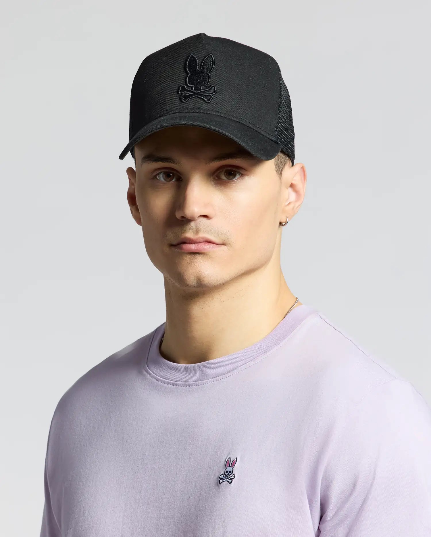 A person wears the MENS WILDER TRUCKER CAP - MO1500026 by Psycho Bunny, featuring a black cap with a bunny skull logo, paired with a light purple shirt bearing the same emblem. The adjustable strap ensures the perfect fit against the plain gray background, combining rugged style and casual charm.