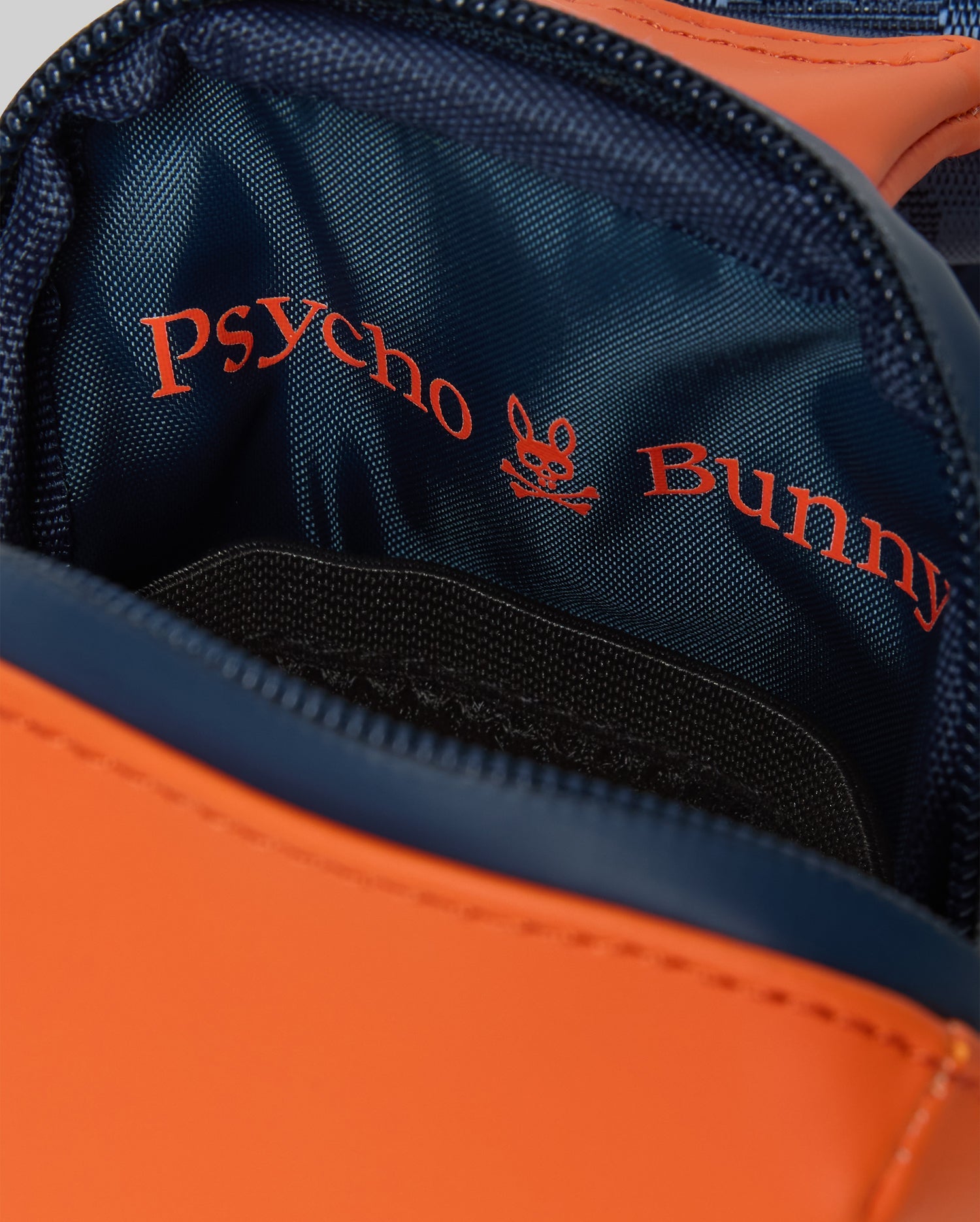 Orange Mens Rubberized Crossbody bag with navy blue trim and interior, featuring a zippered main compartment and a large red Psycho Bunny skull logo and text print on the navy lining.