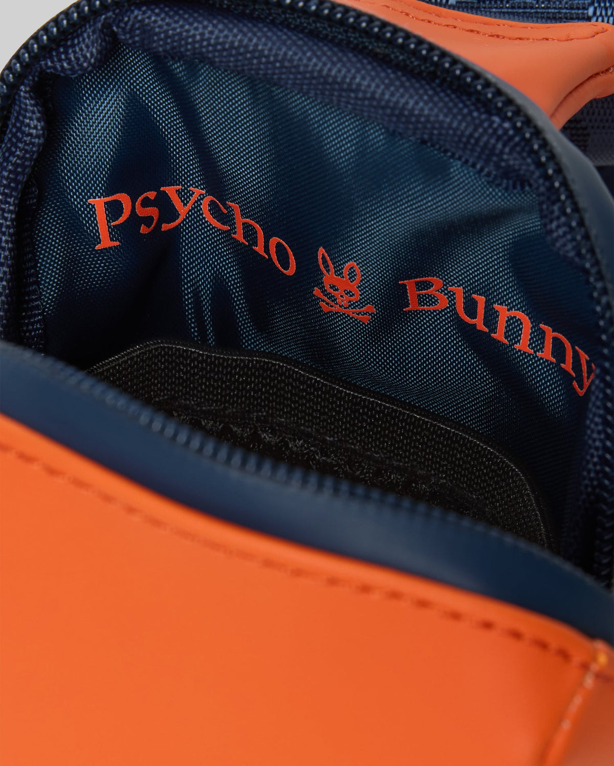 Orange Mens Rubberized Crossbody bag with navy blue trim and interior, featuring a zippered main compartment and a large red Psycho Bunny skull logo and text print on the navy lining.