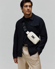 Mens Rubberized Crossbody in cream with a large black Psycho Bunny skull logo, worn with a black collared shirt jacket featuring a small embroidered bunny, a black crew neck tee, and khaki pants.