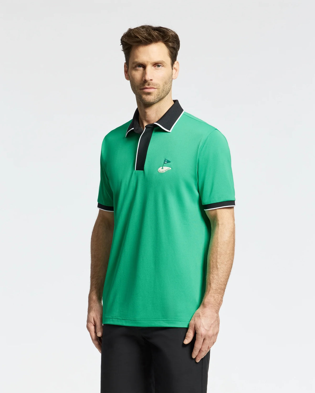 A man wearing the Psycho Bunny MENS CLAYTON PIQUE POLO (MT0100004), designed in a breathable Pima cotton, stands against a plain background. The green shirt is colorblocked with white and black on the collar and sleeves, featuring a black collar and logo on the chest.