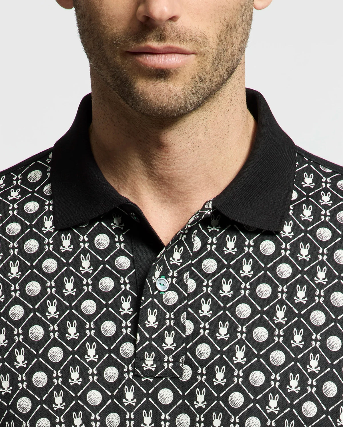 A man is wearing the Psycho Bunny MENS BECKETT PIQUE POLO (MT0100022), made of breathable black Pima cotton with white cartoon bunny faces and circular shapes. Facing the camera, he showcases its distinctive black collar and upper body design.