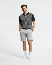 A person stands against a plain background wearing a Psycho Bunny MENS BECKETT PIQUE POLO (MT0100022) in black, light gray shorts, white socks, and white sneakers. With hands in pockets and neatly styled hair, the breathable polo adds an effortless touch to their ensemble.