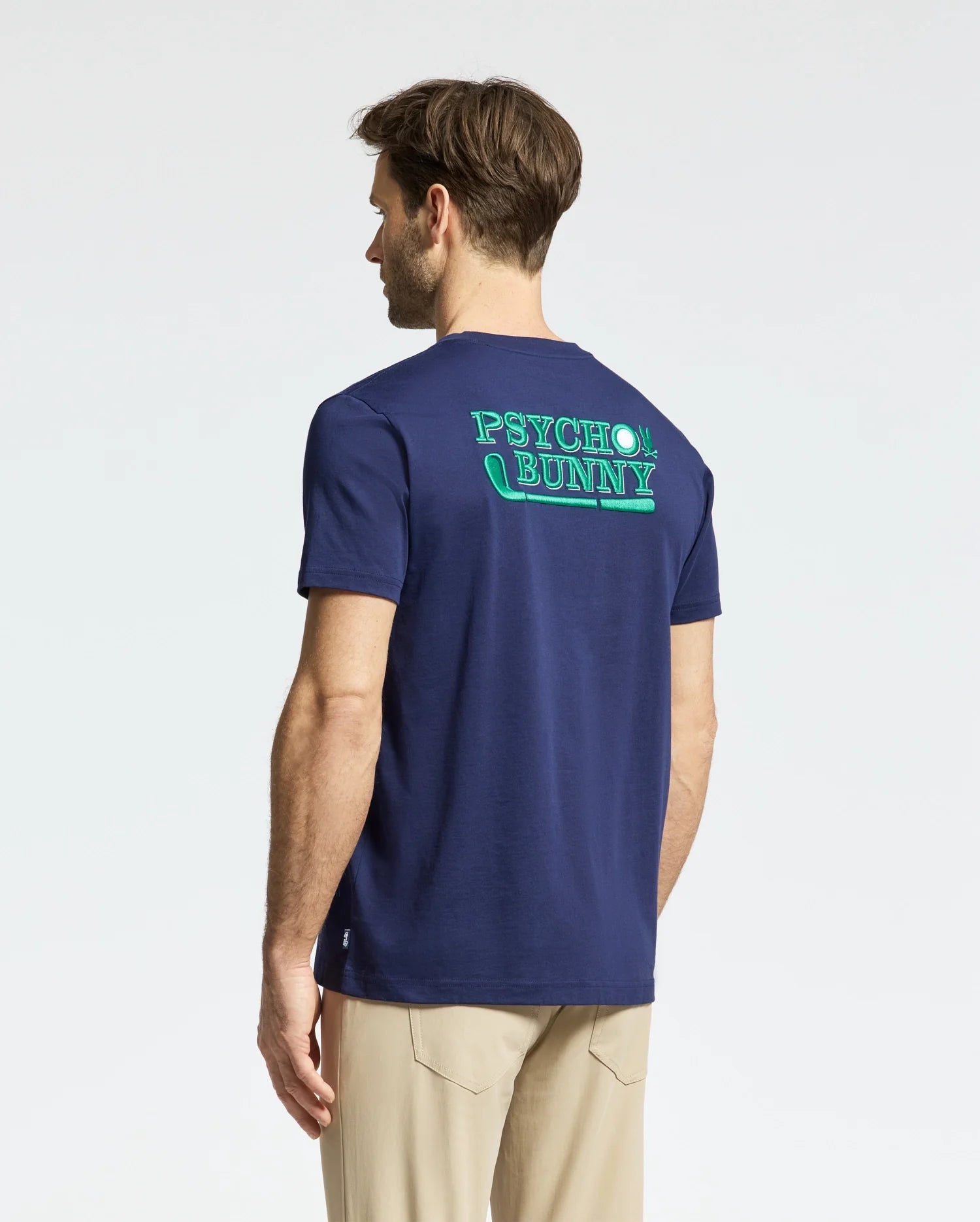 Wearing the MENS DENIS GRAPHIC TEE (MT0200015) by Psycho Bunny, a man showcases a navy Pima cotton T-shirt with bright neon green lettering on the back, pairing it with beige pants. The outfit embodies laid-back elegance with a subtle nod to golf against a simple light backdrop.
