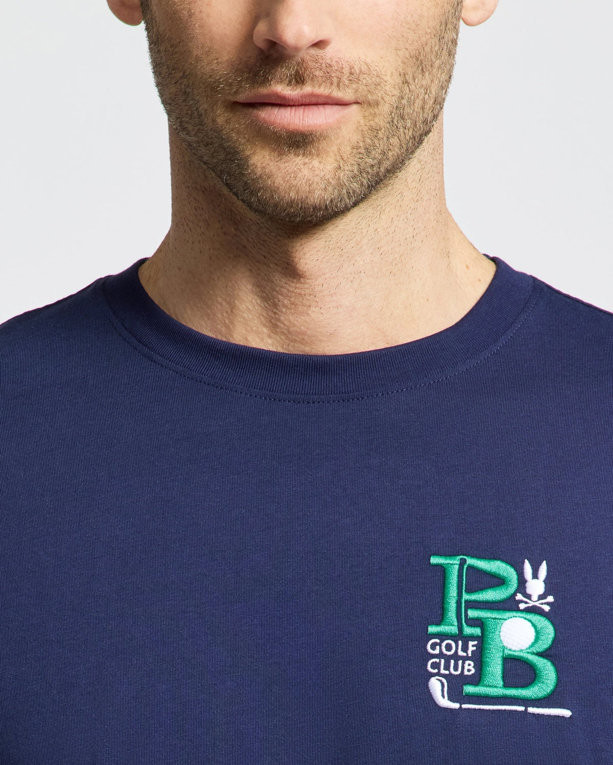 A man dons a Psycho Bunny MENS DENIS GRAPHIC TEE - MT0200015, a navy blue Pima cotton shirt featuring an embroidered PB Golf Club logo on the chest in green and white with golf clubs and a small bird, capturing a golf-inspired aesthetic. Only his upper torso and partial face are visible.