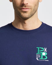 A man dons a Psycho Bunny MENS DENIS GRAPHIC TEE - MT0200015, a navy blue Pima cotton shirt featuring an embroidered PB Golf Club logo on the chest in green and white with golf clubs and a small bird, capturing a golf-inspired aesthetic. Only his upper torso and partial face are visible.