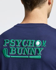 A person wearing the MENS DENIS GRAPHIC TEE - MT0200015 by Psycho Bunny shows "Psycho Bunny" in green embroidery on a navy Pima cotton shirt, featuring an embroidered bunny logo and golf-themed designs.