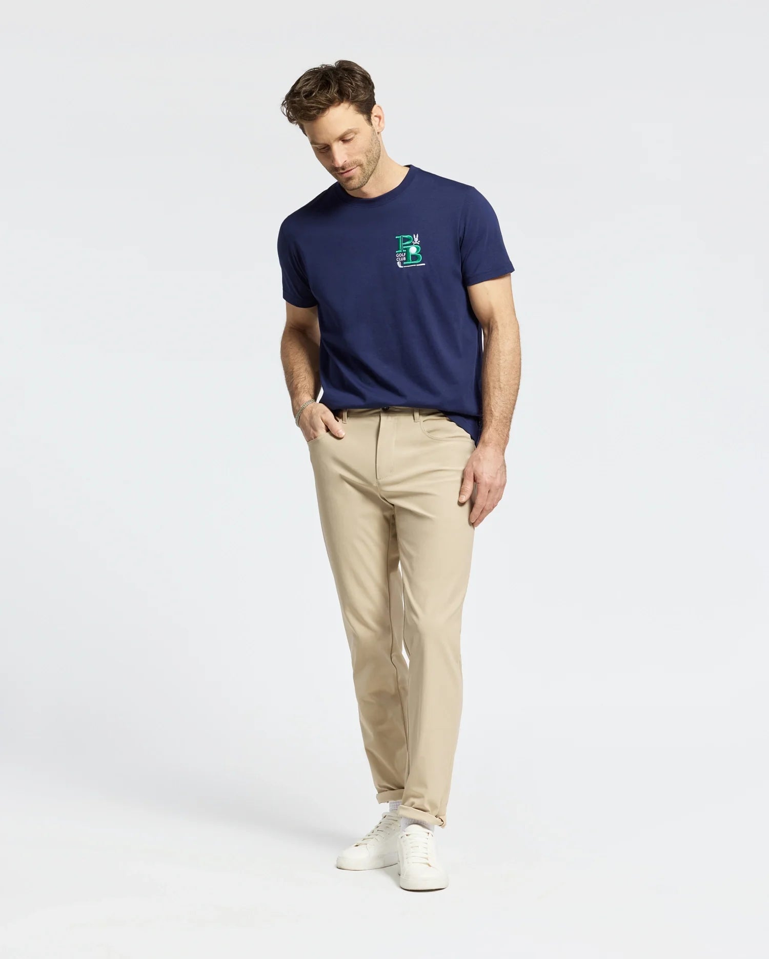 A man is wearing a Psycho Bunny navy MENS DENIS GRAPHIC TEE featuring an embroidered "E" on the chest, paired with beige pants and white sneakers. He stands against a plain white background, looking down with hands in his pockets.