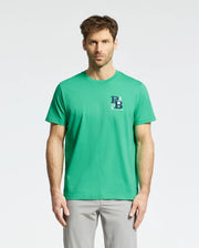 A man wearing a Psycho Bunny Men's Denis Graphic Tee - MT0200015 in green, featuring an embroidered logo on the left side, and light gray pants stands against a plain background.