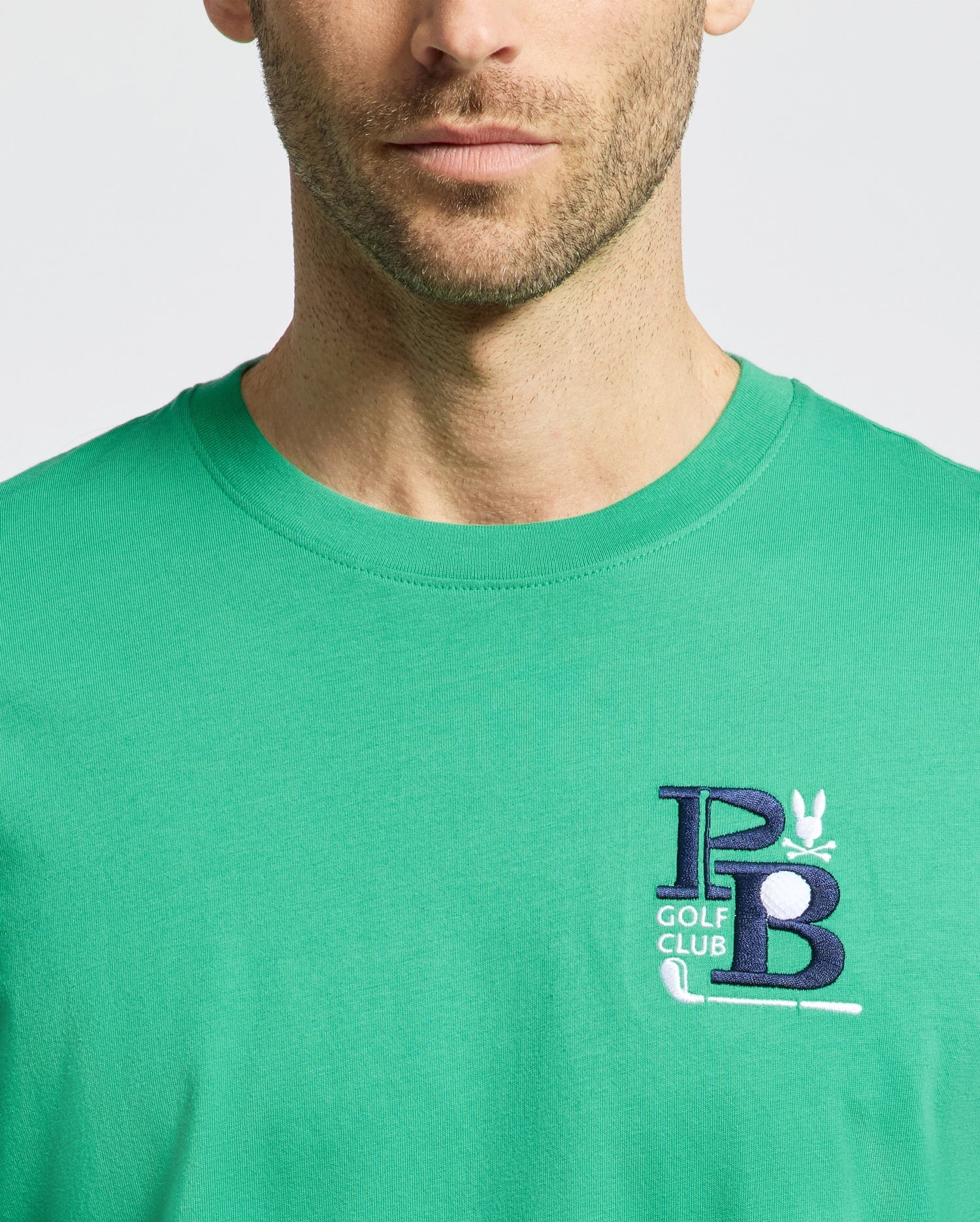A man dons the Psycho Bunny MENS DENIS GRAPHIC TEE - MT0200015 in green Pima cotton, showcasing an embroidered "PB" and "GOLF CLUB" in blue and white on the chest for a golf-inspired style against a plain white backdrop.