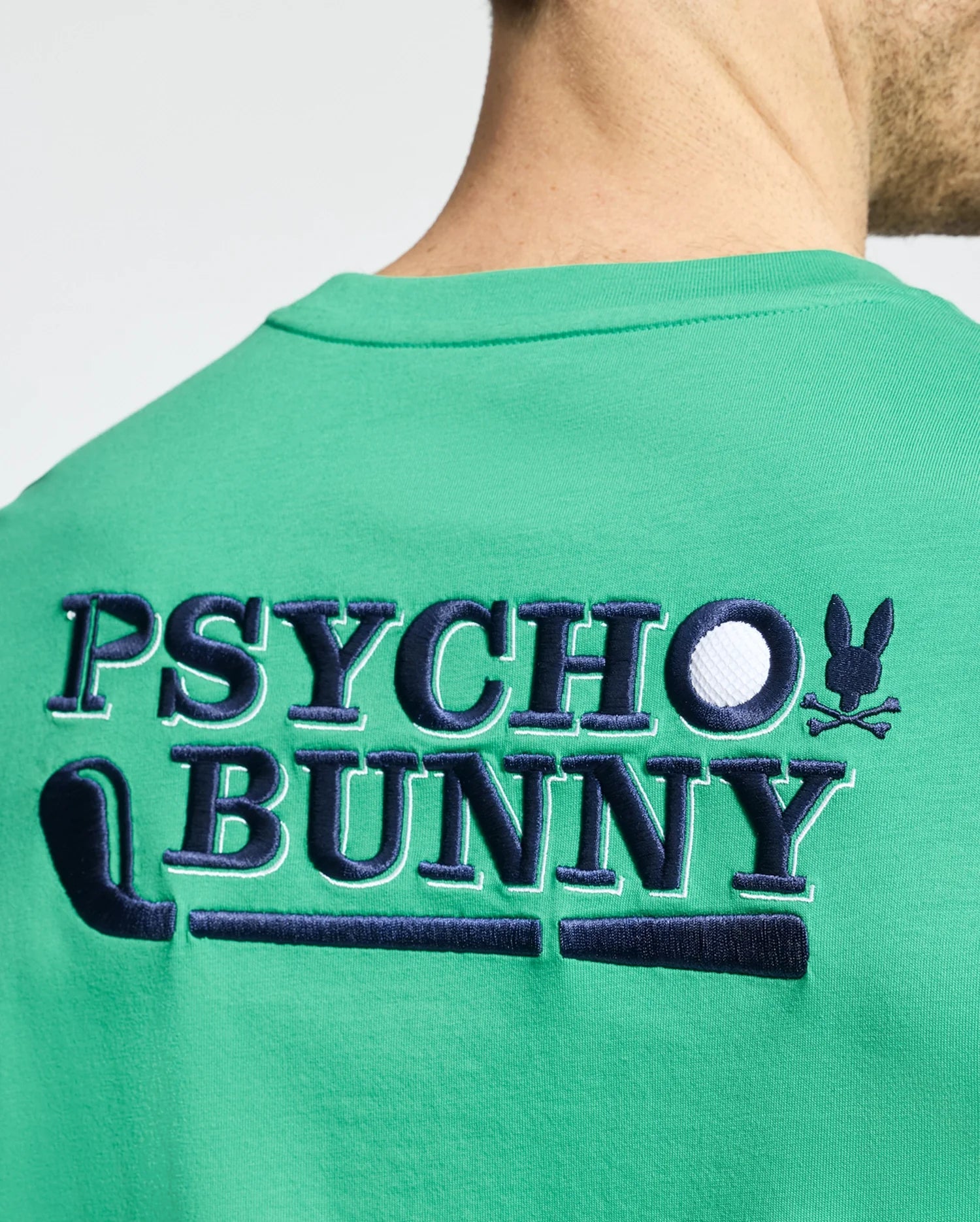 A person wears a MENS DENIS GRAPHIC TEE from Psycho Bunny, crafted from Pima cotton. The green shirt features "Psycho Bunny" embroidered in blue and white on the back, with a golf-inspired yet edgy design, including a small bunny logo with crossbones.