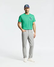 A man stands against a plain background wearing Psycho Bunny's MENS DENIS GRAPHIC TEE (MT0200015) in golf-inspired green, crafted from soft Pima cotton with an embroidered logo, light gray pants, a navy cap featuring a green logo, and white sneakers.